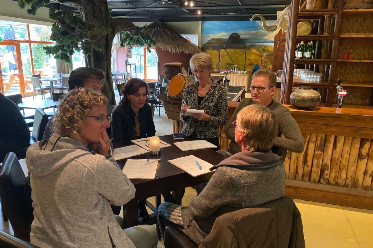 Een groep mensen zit rond een tafel en luistert naar een staande vrouw die leest van een papier in een gezellig ingericht restaurant.