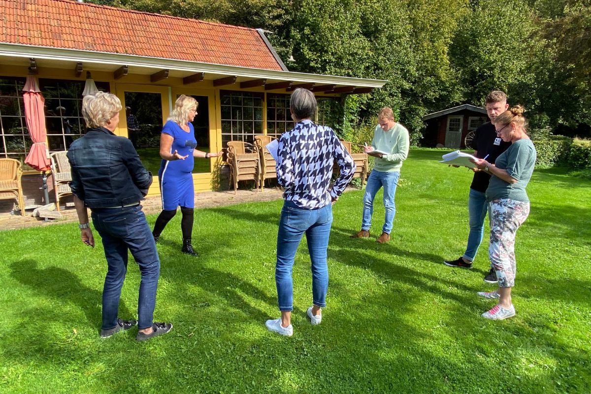 Zeven personen in een discussie buiten bij een huis met natuur op de achtergrond.