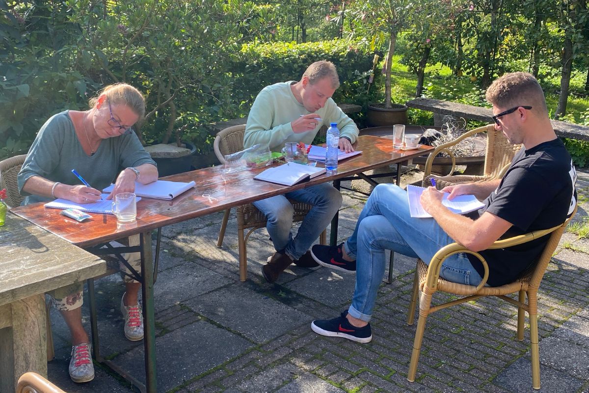 Drie personen zitten aan een buitentafel te schrijven en lezen in een tuinomgeving tijdens een workshop van Big Five for Life.