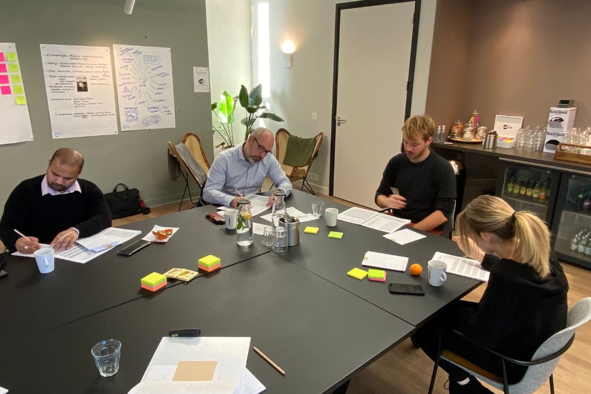 en team van vier personen zit aan een vergadertafel in een moderne kantooromgeving, druk bezig met notities en papieren. Aan de muur hangen flip-over vellen met aantekeningen en diagrammen. Op de tafel liggen kleurrijke post-its, pennen, en drinken. De foto straalt een sfeer van samenwerking en productiviteit uit, passend bij het thema van de blog over het creëren van een positieve teamcultuur.