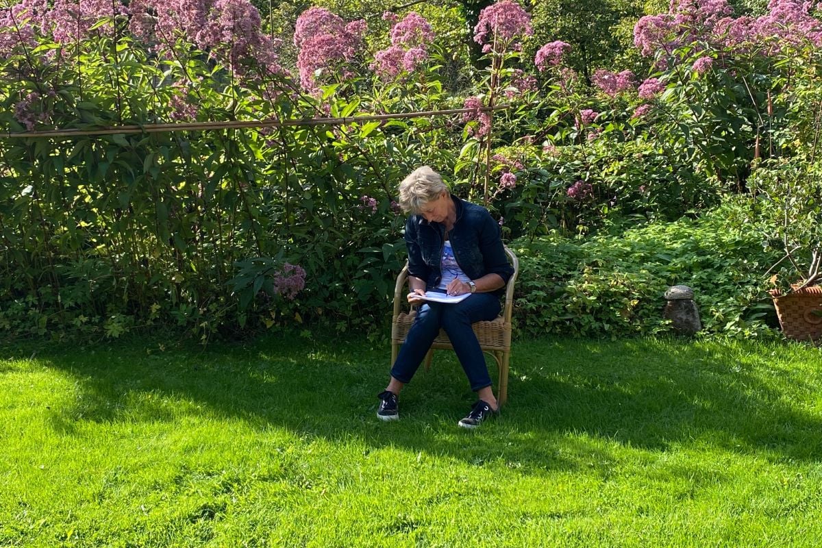 Een vrouw zit in een groene tuin op een stoel en schrijft in een notitieboekje. Ze is omgeven door bloeiende, roze bloemen en de zon schijnt helder. De vrouw neemt de tijd om na te denken en haar gedachten op papier te zetten, symbool voor reflectie en bewuste keuzes maken.