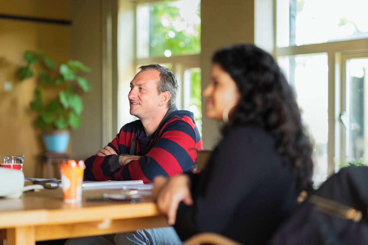 Een man en een vrouw zitten aan een tafel tijdens een bijeenkomst, luisterend en lachend. De man draagt een gestreepte trui en kijkt aandachtig naar voren. De vrouw, gedeeltelijk onscherp, zit naast hem. Op de achtergrond zijn grote ramen en een plant zichtbaar. De afbeelding illustreert werkgeluk en betrokkenheid op de werkplek.