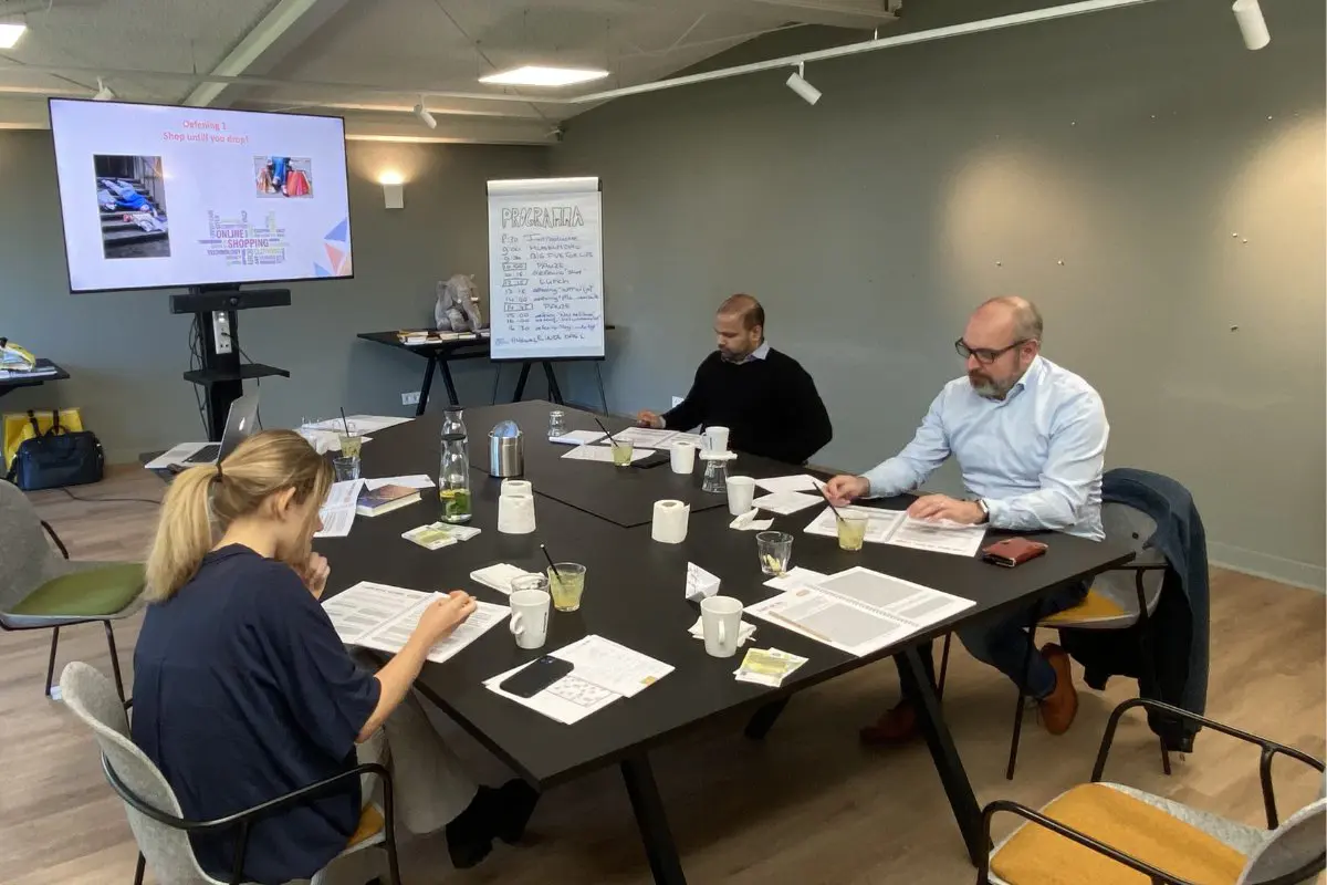 Een kleine groep medewerkers zit aan tafel tijdens een bedrijfstraining. Op de achtergrond is een flip-over met een programma en een scherm met een presentatie over online shoppen zichtbaar. De training is gericht op het bevorderen van transparantie en betrokkenheid binnen het bedrijf, met medewerkers die actief meedoen en aantekeningen maken.