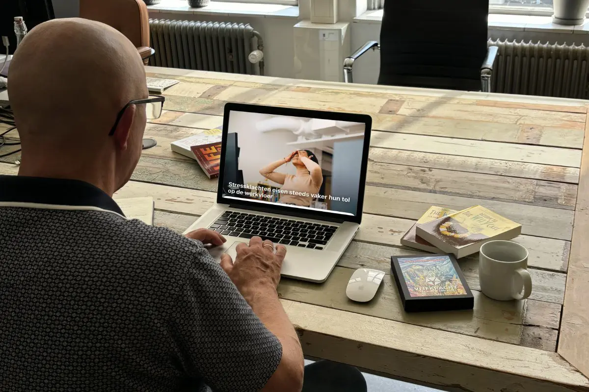 Maurice, Big Five for Life coach, leest een artikel over stressklachten op de werkvloer op een laptop, met boeken en een kop koffie op een houten tafel.