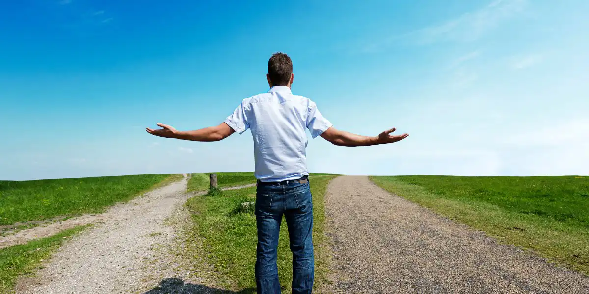 Afbeelding van een man die op een splitsing staat met open armen, symboliserend het maken van keuzes in het leven. Het landschap met twee wegen, groen gras en een blauwe lucht benadrukt het thema van het artikel 'Hoe weet ik of ik op het juiste pad in mijn leven zit.