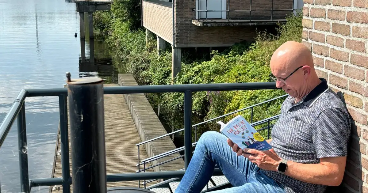 Maurice van Lier leest ontspannen 'The Big Five for Life' op een terras met uitzicht op het water, symbool voor reflectie en inspiratie om jouw mooiste leven te creëren.